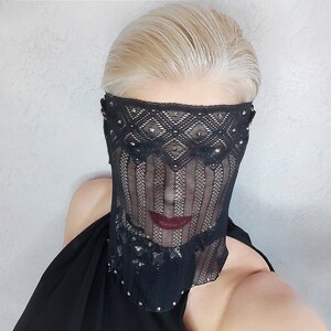 May include: A black lace mask with silver studs covering the face. The mask has a geometric pattern and is worn by a person with blonde hair.
