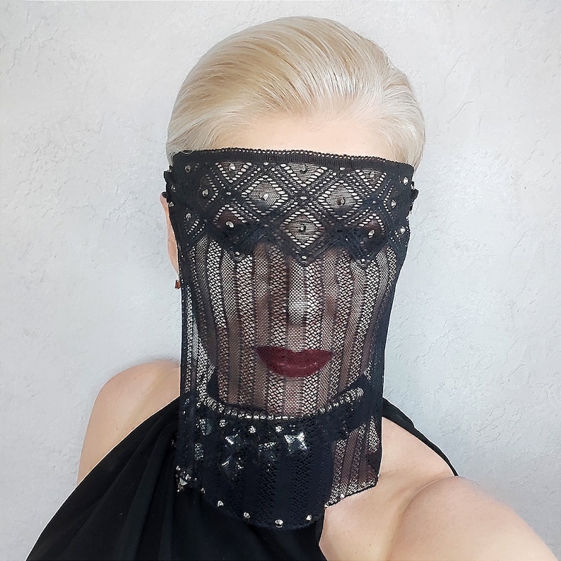 May include: A woman wearing a black lace veil with rhinestones over her face. The veil is sheer and has a geometric pattern. The woman has blonde hair and is wearing a black dress.