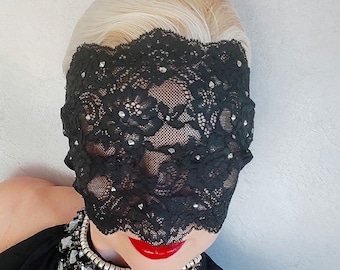 May include: Black lace eye mask with sparkling embellishments. The mask covers the eyes and forehead, with a scalloped edge. The person is wearing a black top and a silver and crystal necklace. Red lipstick completes the look.