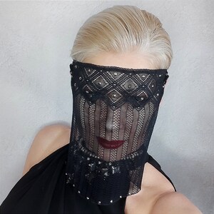 May include: A woman wearing a black lace mask with silver embellishments. The mask covers her eyes and nose, and she is looking directly at the camera.