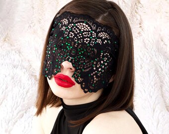 Könnte beinhalten: Eine schwarze Spitzen-Augenmaske mit grünen Akzenten bedeckt die Augen. Die Person trägt roten Lippenstift und ein schwarzes Oberteil. Der Hintergrund ist ein weiches, weißes, strukturiertes Gewebe.