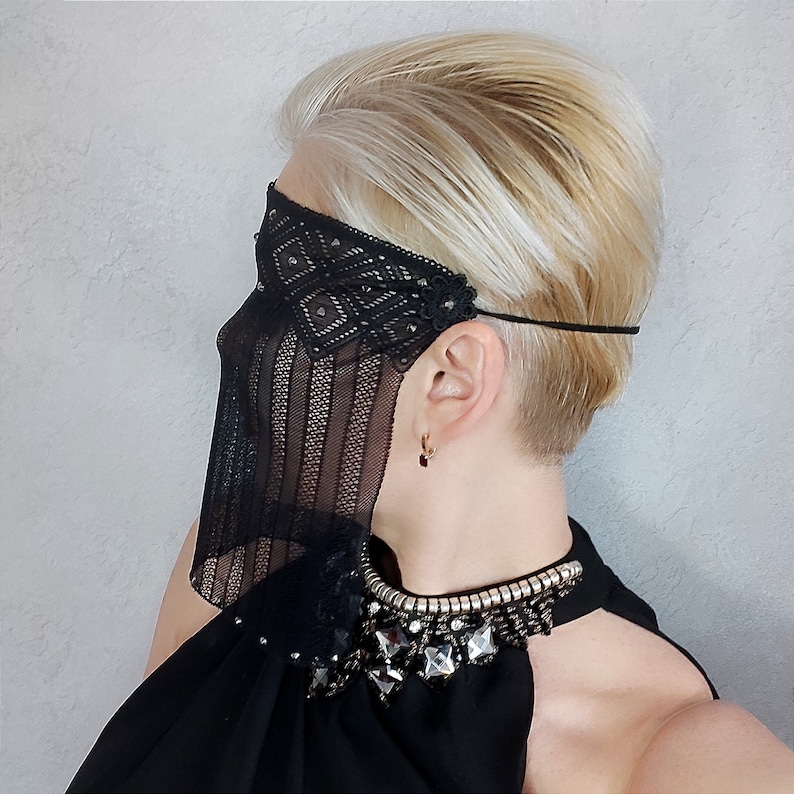 May include: A black lace mask with rhinestones covers the eyes of a person with short blonde hair. The mask is attached to a black headband. The person is wearing a black top with a beaded neckline.