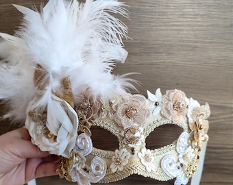 Op de afbeelding: Een sierlijk, crèmekleurig masker voor een gemaskerd bal met gouden en witte versieringen. Het masker heeft delicate bloemenapplicaties, kralen en kanten details. Een tros witte veren en een stoffen bloem sieren de linkerkant.