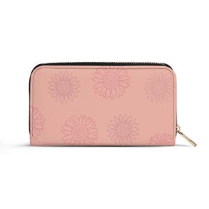 May include: A blush pink wallet with a black zip and a gold-coloured zip pull. The wallet features a repeating pattern of light pink sunflowers.