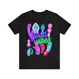 May include: A black short-sleeve t-shirt featuring a graphic print of various colorful, cartoon-style adult toys with smiling faces. The text "Good vibes" is written in a neon-style script in the center, with purple and green glowing accents.