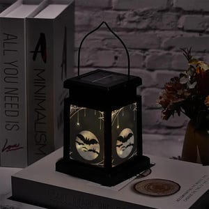 May include: Black solar-powered lantern with a handle, featuring Halloween designs. The lantern has a square shape with panels displaying bats and spiderwebs. The lantern is sitting on a book.