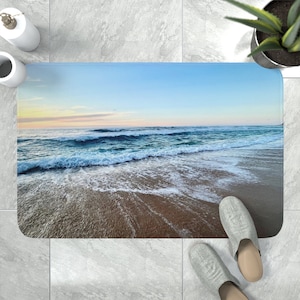 May include: A rectangular bath mat featuring a high-resolution photographic print of an ocean shoreline at sunset. The image displays blue waves with white foam crashing onto a sandy beach under a soft, pale yellow and light blue sky.