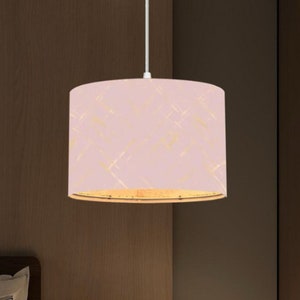 May include: A pink pendant light with a cylindrical lampshade featuring a gold abstract pattern. The light hangs from a white cord. The background is a dark brown wall. The light is suitable for a bedroom.