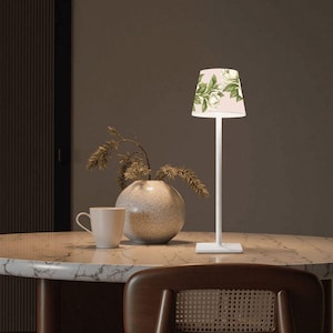 May include: A tall, slender white table lamp with a tapered shade featuring a light pink background and a botanical print of white flowers and green leaves. The lamp has a square base and a thin, straight vertical stem that supports the illuminated shade.