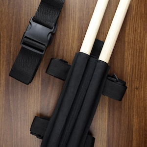 May include: A black nylon holster with two vertical slots containing light-coloured wooden dowels. The holster features adjustable black webbing straps with plastic side-release buckles, designed for secure attachment and carrying of the wooden items.