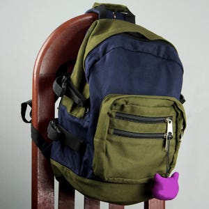 Puede incluir: Una mochila de dos tonos con paneles de tela en verde oliva y azul marino. La bolsa incluye un bolsillo frontal con dos compartimentos horizontales con cremallera, correas laterales negras ajustables y un pequeño dije morado con forma de gato.