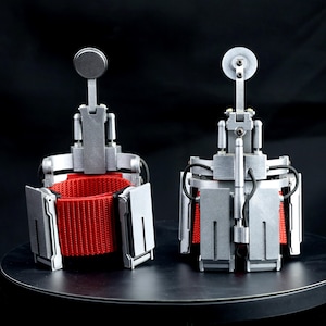 May include: Two silver-colored mechanical wrist-mounted devices featuring a bright red fabric strap interior. Each unit has a rigid metallic-looking frame with articulated components, a vertical post with a circular top, and black wiring details.