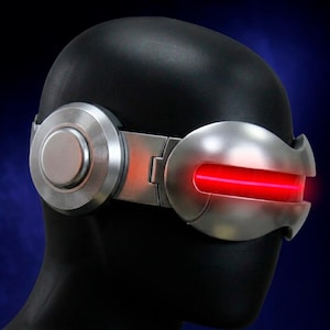 May include: A silver-coloured visor with a horizontal red light strip across the front, featuring circular side panels with concentric ring detailing. The visor is positioned on a black mannequin head against a dark blue background.