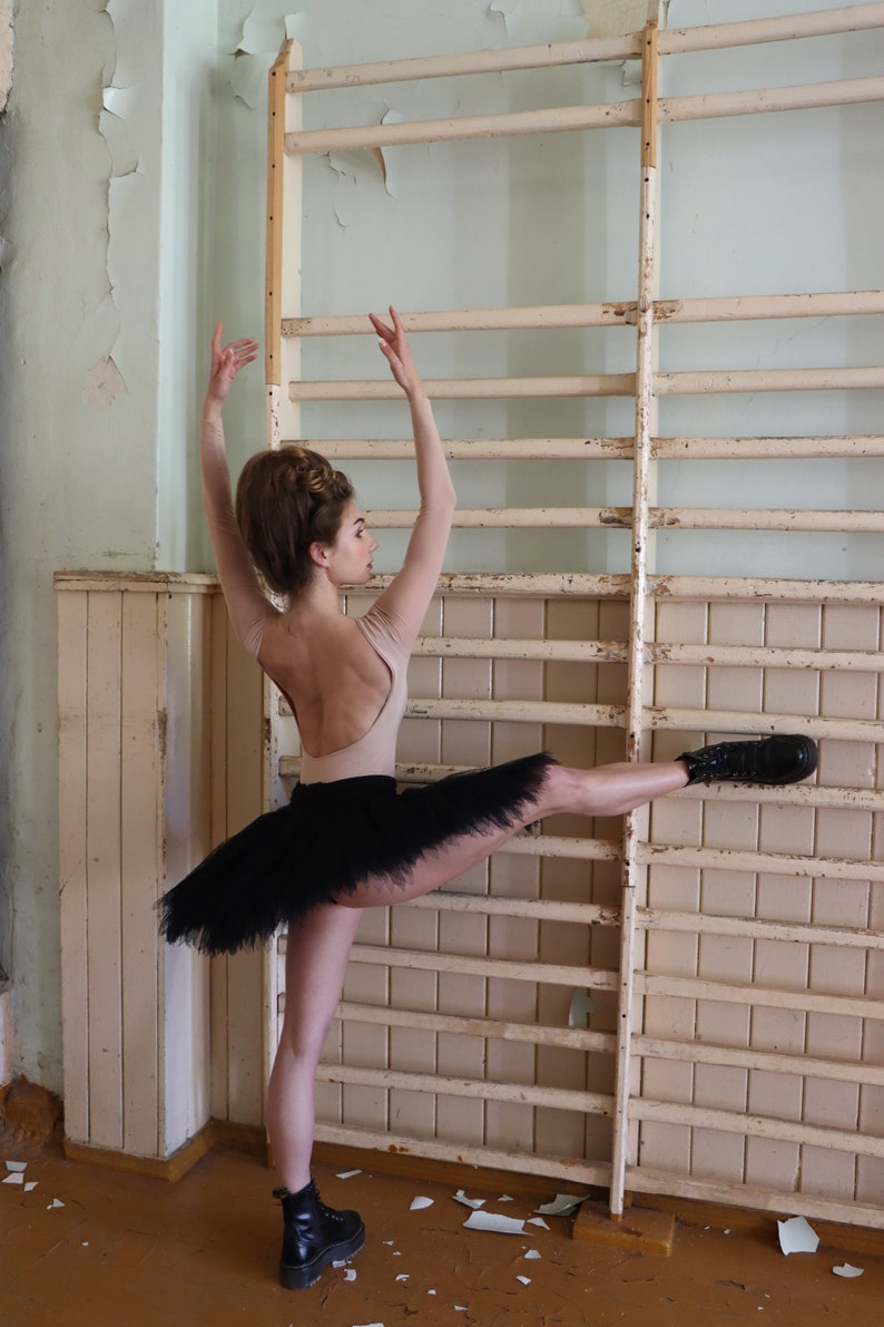 K&ouml;nnte beinhalten: Eine Ballerina in einem schwarzen Tutu und einem beigen Trikot f&uuml;hrt eine Bein-Extension-Pose vor einer Holzwand mit einer wei&szlig; gestrichenen Leiter aus. Die Ballerina tr&auml;gt schwarze Stiefel.
