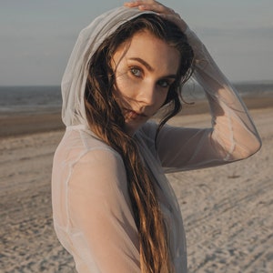 May include: A woman with long brown hair wearing a white sheer hooded top. She is standing on a sandy beach with the sea in the background.
