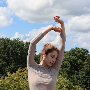 K&ouml;nnte beinhalten: Eine Frau tr&auml;gt ein durchsichtiges, hellgraues Langarmshirt mit hoch erhobenen Armen. Sie steht im Freien vor einem Hintergrund aus gr&uuml;nen B&auml;umen und einem blauen Himmel mit wei&szlig;en Wolken.