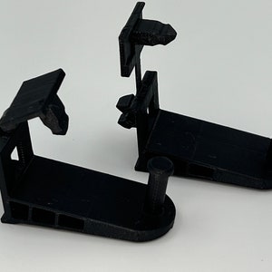 Può includere: Due ganci da parete in plastica nera con una base arrotondata e un piccolo gancio nella parte superiore. I ganci sono progettati per essere montati su una parete.