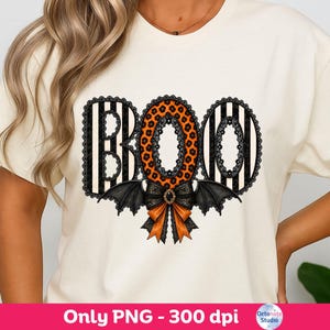 以下が含まれることがあります： クリーム色のTシャツで、BOOという文字のグラフィックが描かれています。BとOの文字は黒と白のストライプで、中央のOはオレンジ色のヒョウ柄です。デザインには黒いレースの縁取り、コウモリの羽、リボンが含まれています。