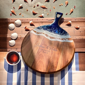 May include: A round wooden serving board with a blue and white epoxy resin design resembling ocean waves. The board has a handle and is engraved with the text "Joshua & Heller - ENGAGED - 12.16.24".
