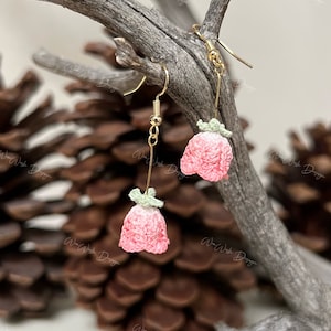 May include: A pair of pink and green crocheted flower earrings with gold hooks. The earrings are hanging from a branch.