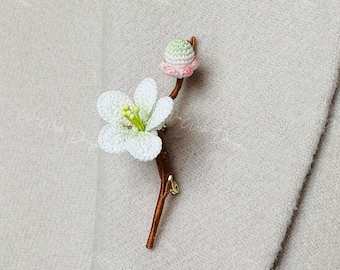 May include: A white crocheted flower with a green and pink bud on a brown stem. The flower is attached to a pin, making it a brooch.