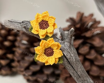 May include: Two handmade crochet sunflower earrings. The earrings feature bright yellow petals with a brown center and green leaves. The earrings are displayed on a piece of driftwood.