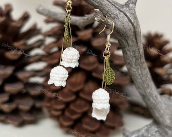 May include: Handmade white crochet lily of the valley earrings with gold-colored hooks. The earrings feature delicate white flower buds and green leaves, suspended from gold-tone wire. The earrings are displayed on a natural wooden branch against a blurred background.