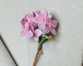 May include: A delicate crocheted hydrangea flower brooch in shades of pink and purple with a green stem and a gold pin.