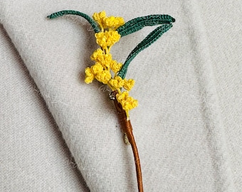 May include: A handmade crochet brooch featuring a sprig of yellow mimosa flowers with green leaves. The stem is wrapped in brown thread. The brooch is set against a light gray fabric background.