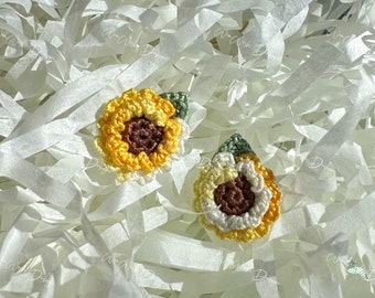 May include: Two crocheted sunflower embellishments with brown centers, yellow petals, and green leaves. The sunflowers are on a white background.