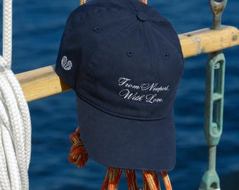 May include: A navy blue baseball cap featuring white embroidered text on the front that reads From Newport, With Love. The side of the cap displays a small white embroidered icon of two seashells. The cap has a structured crown and a curved brim.
