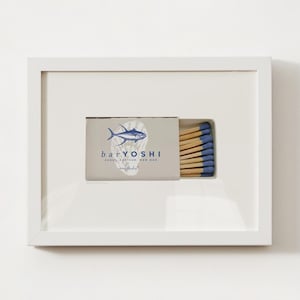Op de afbeelding: Een witgelijste print met een lucifersdoosje met de tekst "bar YOSHI" en een blauwe visillustratie. Het lucifersdoosje bevat houten lucifers met blauwe uiteinden. De print staat op een witte achtergrond.