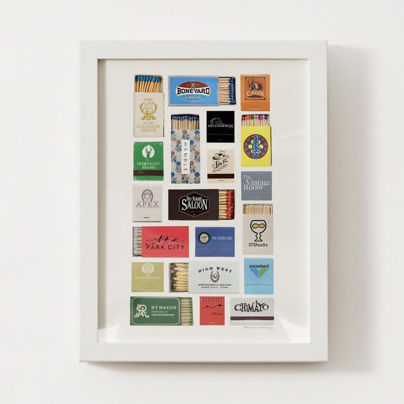May include: A white frame containing a collection of matchboxes. The matchboxes are arranged in a grid pattern and feature various designs and logos, including "Boneyard", "Cabin", "Handle", "No Name Saloon", "Park City", "O'Shucks", "Snowbird", "St. Regis", and "Chimayo".