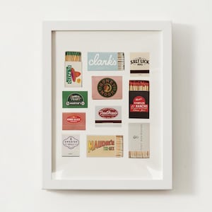 May include: A white frame with a collection of vintage matchboxes. The matchboxes feature various designs and logos, including "El Patio", "Martin's", "Home Slice Pizza", "Clark's", "Honey Moon", "Food Heads", "Swedish Mill", "Maudie's", "Salt Lick BBQ", "Matt's Famous El Rancho", and "Tiny Boxwoods".