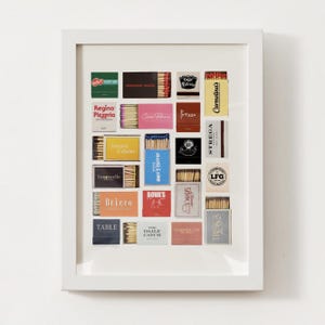 Op de afbeelding: Een wit frame met een collage van luciferdoosjes met verschillende restaurant- en cafénamen. De luciferdoosjes zijn in een rasterpatroon gerangschikt en bevatten namen zoals "Regina Pizzeria", "Mamma Maria", "Cine Prezza", "Stregga", "Bricco", "The Daily Catch" en "Limoncello".