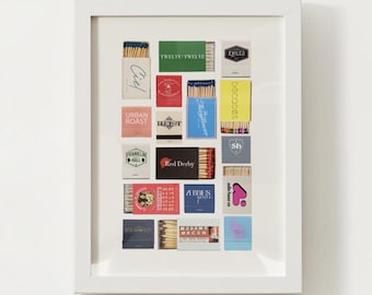 May include: A white frame with a collection of matchboxes inside. The matchboxes are arranged in a grid pattern and feature various designs and logos, including "Exiles", "Twelve Twelve", "Ciel", "Urban Roast", "Franklin Hall", "Decoodes", "Mayflower", "Sh", "Red Derby", "Abbies", "Aslin Beer Co", and "Madams Organ".