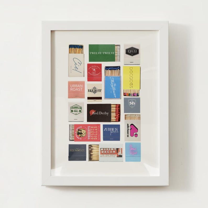 May include: A white frame with a collection of matchboxes inside. The matchboxes are arranged in a grid pattern and feature various designs and logos, including "Exiles", "Twelve Twelve", "Ciel", "Urban Roast", "Franklin Hall", "Decoodes", "Mayflower", "Sh", "Red Derby", "Abbies", "Aslin Beer Co", and "Madams Organ".