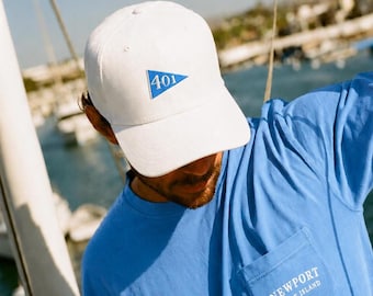 May include: White baseball cap with a blue triangle emblem featuring the number "401". A blue t-shirt with a pocket that reads "Newport Rhode Island". The image is taken outdoors.