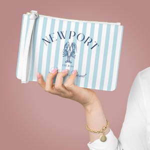 Op de afbeelding: Een clutch met verticale witte en lichtblauwe strepen, met het woord "NEWPORT" in marineblauw. Het heeft een marineblauwe kreeftafbeelding en de woorden "Sea Sand" eronder. Het heeft een witte polsband en een gouden rits.