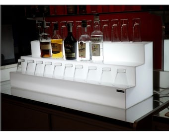 Pode incluir: Prateleira de acrílico branca de dois níveis com luz LED integrada, que exibe garrafas de licor e copos de shot.