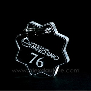 Può includere: Un portachiavi a forma di stella in acrilico trasparente con un anello argentato. Il portachiavi ha il testo "Hotel Ristorante Marechiaro" e il numero "76" stampato su di esso. Il testo "www.plexdautre.com" è stampato sul fondo del portachiavi.