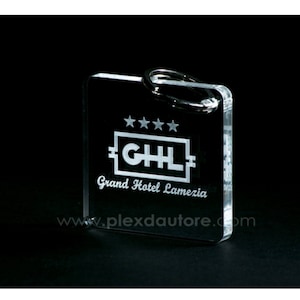Può includere: Un portachiavi in acrilico trasparente con un logo inciso in bianco che dice "Grand Hotel Lamezia" e "GHL" con quattro stelle sopra il logo. Il sito web "www.plexdaautore.com" è anche inciso sul portachiavi.
