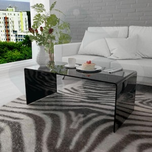 以下が含まれることがあります： 透明な天板と湾曲したデザインの黒いガラス製のコーヒーテーブル。テーブルはリビングルームのゼブラ柄の敷物の上に置かれています。