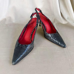May include: A pair of grey snakeskin-effect slingback heels with pointed toes. The interior is bright red, and the heels are thin. The shoes have a buckle closure. The shoes are set against a neutral background.