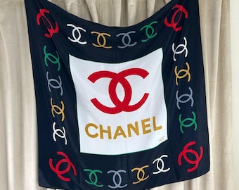 May include: Black square scarf with a white center featuring a red double-C logo and the word "CHANEL" in gold. The scarf has a border of colorful interlocking double-C logos.