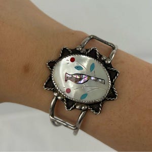 May include: Silver cuff bracelet with a central oval design featuring a bird inlay. The bird is made of iridescent material, with red and turquoise accents. The bracelet has a sunburst-like border.