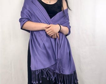 May include: A periwinkle-colored shawl with fringe. The shawl is draped over a black top and dress. The fringe hangs at the bottom of the shawl. The shawl is made of a soft, flowing fabric.