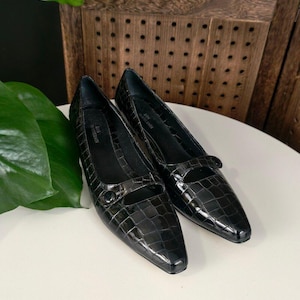May include: A pair of black patent leather shoes with a glossy crocodile-embossed texture. These shoes feature a pointed toe, a low block heel, and a decorative strap across the vamp secured with a small circular button. The interior is black.