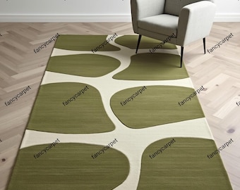 Op de afbeelding: Een rechthoekig vloerkleed met een modern design. Het kleed heeft olijfgroene organische vormen op een gebroken witte achtergrond. Een lichtgrijze fauteuil met zwarte poten staat op het kleed, dat op een houten vloer ligt.