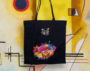 May include: Black canvas tote bag with a colorful graphic of a pair of lips with butterflies and paint splatters. The bag has two black straps.